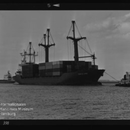 Containerschiff Carolina mit drei Ladekränen und aufgetürmten Containern, begleitet von Schleppern im Hafen.