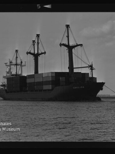Containerschiff Carolina mit drei Ladekränen und aufgetürmten Containern, begleitet von Schleppern im Hafen.