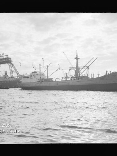 Frachtschiff im Hafen vor großen Krananlagen und Werft, ruhige See im Vordergrund.