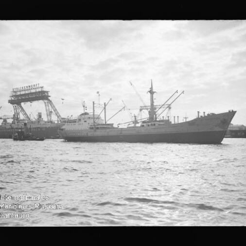 Frachtschiff im Hafen vor großen Krananlagen und Werft, ruhige See im Vordergrund.