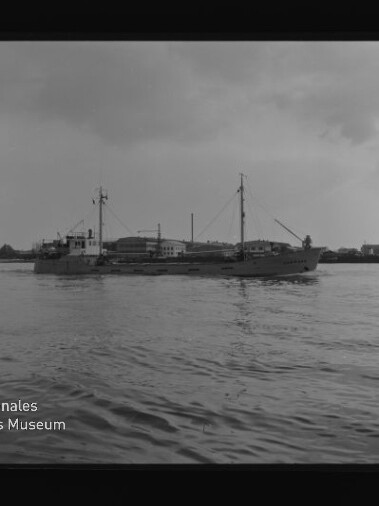 Schwarzweiß-Foto eines Frachtschiffs auf ruhigem Wasser vor Hafenbauten; Wasserzeichen 'Internationales Maritimes Museum Hamburg'.