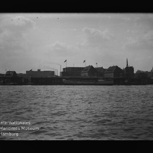 Blick über Wasser auf Hafenfront mit Gebäuden, mehreren Fahnen und Fabrikschornsteinen am Ufer.