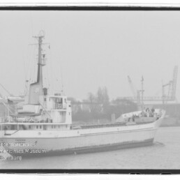 Schwarzweißaufnahme eines Frachtschiffs im Hafen mit Kranen und Industrieanlagen im Hintergrund, leicht neblige Atmosphäre und Museums-Wasserzeichen.