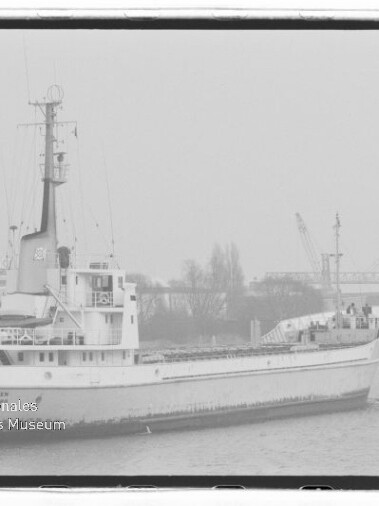 Schwarzweißaufnahme eines Frachtschiffs im Hafen mit Kranen und Industrieanlagen im Hintergrund, leicht neblige Atmosphäre und Museums-Wasserzeichen.