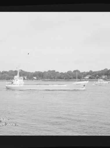 Frachtschiff fährt auf ruhigem Fluss vor baumbewachsener Uferlinie mit kleinem Ausflugsschiff und Museumswasserzeichen.
