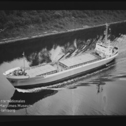 Schwarzweißfoto: Frachtschiff mit abgedeckten Laderäumen fährt einen Kanal entlang; Uferbäume und Museumslogo sichtbar.