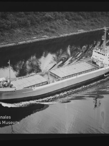 Schwarzweißfoto: Frachtschiff mit abgedeckten Laderäumen fährt einen Kanal entlang; Uferbäume und Museumslogo sichtbar.