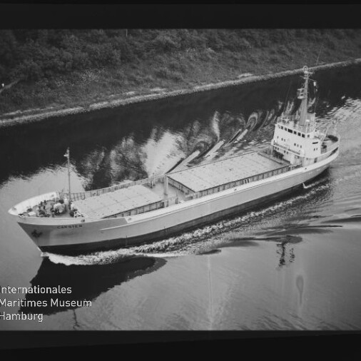 Schwarzweißfoto: Frachtschiff mit abgedeckten Laderäumen fährt einen Kanal entlang; Uferbäume und Museumslogo sichtbar.