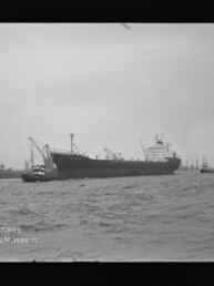 Großes Frachtschiff im Hafen, begleitet von Schleppern, mit Krananlagen und weiteren Schiffen im Hintergrund.