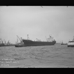 Großes Frachtschiff im Hafen, begleitet von Schleppern, mit Krananlagen und weiteren Schiffen im Hintergrund.