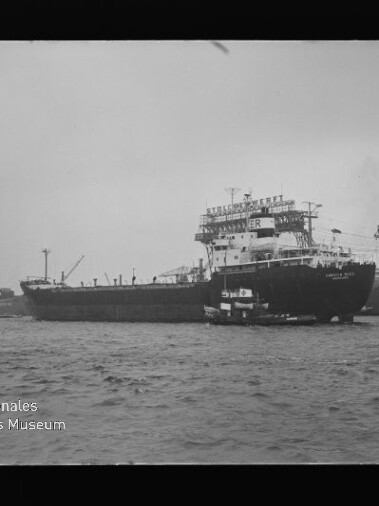 Großes Frachtschiff mit Schlepper neben Bug im Hafen, Kräne im Hintergrund und Museumslogo unten links.