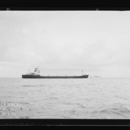 Schwarzweißaufnahme eines Frachtschiffs auf ruhiger See, kleines Boot in der Ferne, Holzpfahl rechts; Wasserzeichen des Internationalen Maritimen Museums Hamburg.