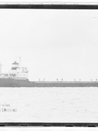 Schwarzweiß-Seitenansicht eines Frachtschiffs mit langem Rumpf und mittschiffs liegender Aufbauten auf ruhiger See.