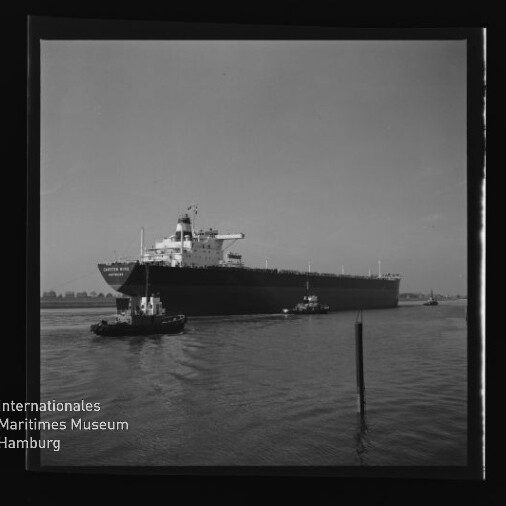 Großes Tankschiff auf dem Wasser, von zwei Schleppern begleitet; Internationales Maritimes Museum Hamburg-Logo unten links.