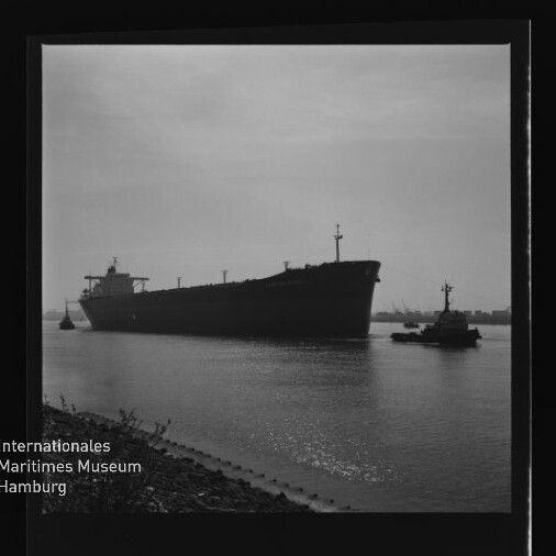 Großes Frachtschiff in Silhouette auf ruhigem Wasser, begleitet von einem Schlepper, mit Museumssignet unten links.