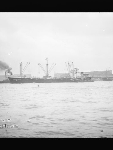Frachtschiff mit rauchendem Schornstein und Ladekränen vor Hafenanlagen, Wasser im Vordergrund, Wasserzeichen unten links.