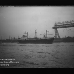 Frachtschiff vor Industriehafen mit großem Portalkran und sichtbarem Schriftzug und Museumslogo unten links.