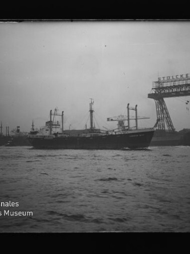 Frachtschiff vor Industriehafen mit großem Portalkran und sichtbarem Schriftzug und Museumslogo unten links.
