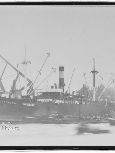Großes Frachtschiff mit Schornstein und Ladekränen am Hafen, Kranbühne und Gebäude im Hintergrund.