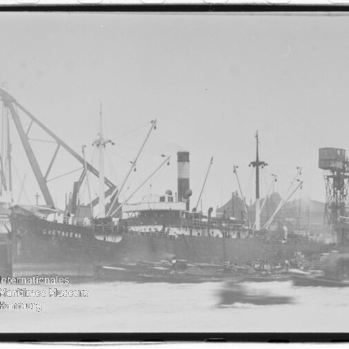 Großes Frachtschiff mit Schornstein und Ladekränen am Hafen, Kranbühne und Gebäude im Hintergrund.
