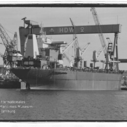 Großes Frachtschiff am Werftkai vor mehreren Hafenkränen und einem Portalkran mit HDW‑Schriftzug, Wasser im Vordergrund.