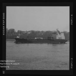 Schwarzweißbild eines Frachtschiffs auf ruhiger See vor bewaldetem Ufer; Filmrand mit KODAK-Markierung und Museumslogo.