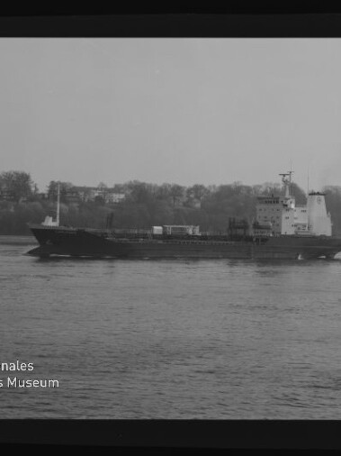 Schwarzweißbild eines Frachtschiffs auf ruhiger See vor bewaldetem Ufer; Filmrand mit KODAK-Markierung und Museumslogo.