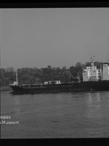 Schwarzweißfoto eines Frachtschiffs auf ruhigem Wasser vor bewaldetem Ufer, mit Filmrand (KODAK FXP 6040) und Museumslogo.