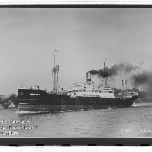 Dampfschiff mit rauchendem Schornstein fährt nahe Küste; links unten Wasserzeichen Internationales Maritimes Museum Hamburg.