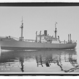 Großes Frachtschiff mit Schornstein und Masten fährt auf spiegelglattem Meer, Reflexion sichtbar.