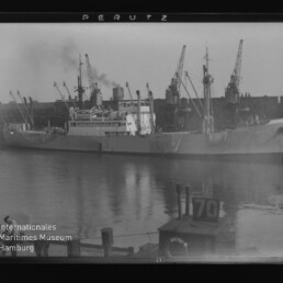 Großes Schiff liegt im Hafen neben Kränen, Spiegelung im Wasser; Schwarzweißaufnahme mit 