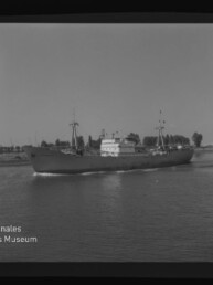 Schwarzweißaufnahme eines Frachtschiffs mit zwei Masten auf ruhigem Wasser, Ufer mit Bäumen im Hintergrund und Museumslogo sichtbar.
