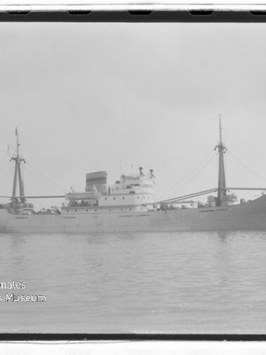 Graustufenaufnahme eines Frachters mit Mittelschiffsaufbau, zwei Masten und Schornstein auf ruhigem Wasser; Museumslogo links unten.
