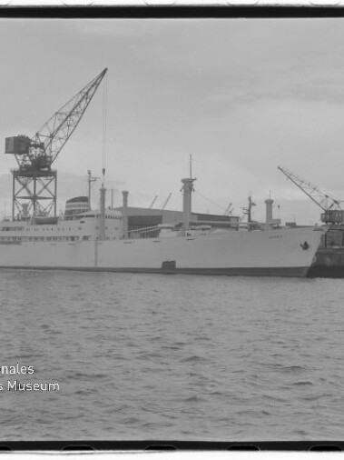 Schwarzweißfoto eines weißen Frachtschiffs am Kai mit großen Hafenkränen und Wasser im Vordergrund; Stempel: Internationales Maritimes Museum Hamburg