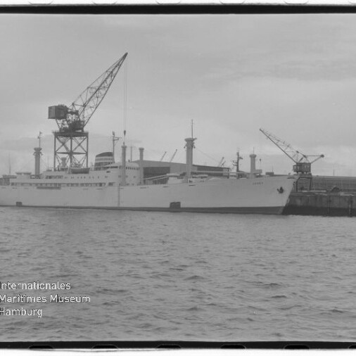 Schwarzweißfoto eines weißen Frachtschiffs am Kai mit großen Hafenkränen und Wasser im Vordergrund; Stempel: Internationales Maritimes Museum Hamburg