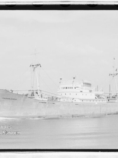 Schwarzweißfoto des Frachtschiffs CASABLANCA mit Ladekränen auf ruhigem Wasser vor der Küste; Museumssiegel unten links.