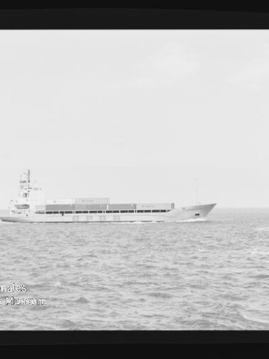Schwarzweißaufnahme eines niedrigen Frachtschiffs mit Containern auf ruhiger See, seitliche Ansicht mit Filmrand und Museumslogo.