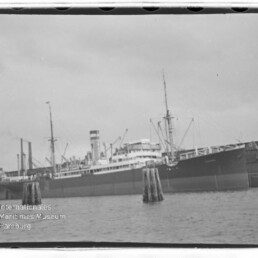 Großes Dampfschiff mit Schornstein und Masten am Kai, Pfähle im Vordergrund; Wasserzeichen Internationales Maritimes Museum Hamburg.