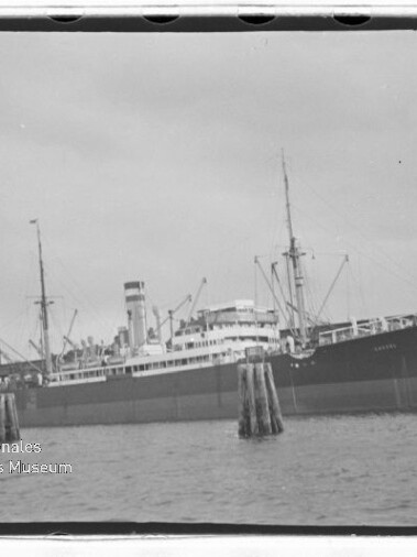 Großes Dampfschiff mit Schornstein und Masten am Kai, Pfähle im Vordergrund; Wasserzeichen Internationales Maritimes Museum Hamburg.