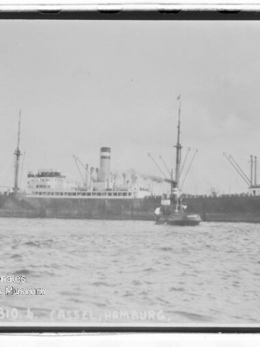 Großes Dampfschiff mit Schornstein und Ladekränen, ein Schlepper an der Seite auf ruhiger See in einer historischen Schwarzweißaufnahme.