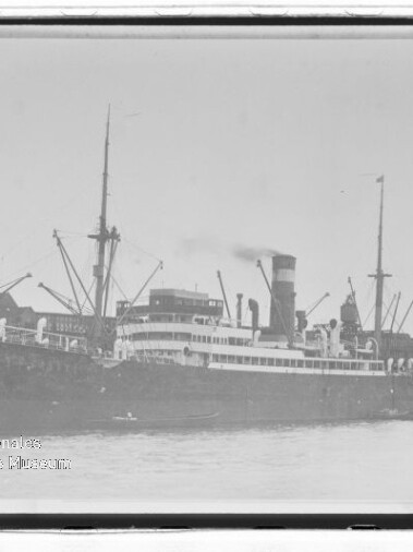 Großes Dampfschiff mit hohem Schornstein und Ladekränen am Kai, Ufergebäude im Hintergrund und ruhige Wasserfläche.
