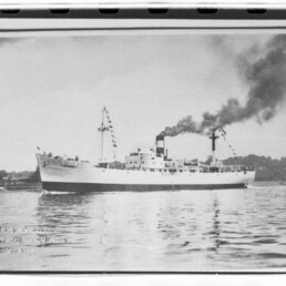 Schwarzweißfoto eines Dampfschiffs, beflaggt, mit rauchendem Schornstein und Spiegelung im ruhigen Wasser.