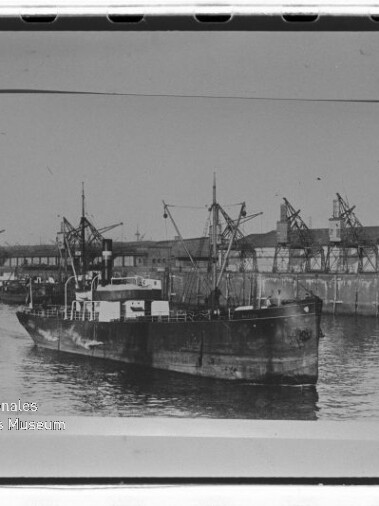 Frachtschiff im Hafen vor Lagerhäusern und Kränen in Schwarzweiß, mit Wasserzeichen Internationales Maritimes Museum Hamburg.