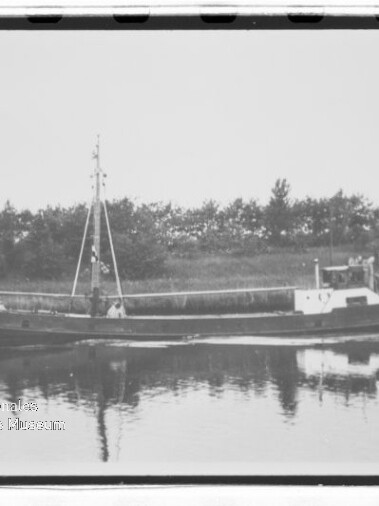Kleines Frachtschiff mit Mast auf ruhigem Wasser, Spiegelung und Baumreihe im Hintergrund; Museumswasserzeichen.