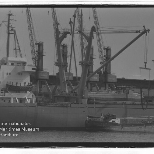 Frachtschiff am Hafen mit mehreren Ladekränen und einer kleinen Barkasse, schwarzweißes Bild mit Museumslogo unten links.