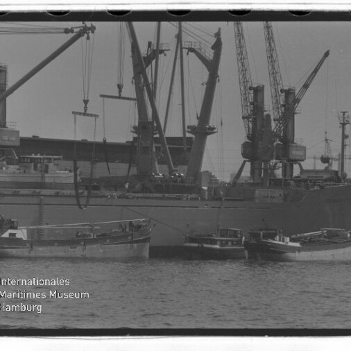 Frachtschiff mit mehreren Ladekränen am Kai und kleinen Booten im Vordergrund; Wasserzeichen des Internationalen Maritimen Museums Hamburg.