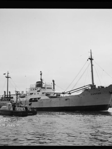 Schwarzweiß-Foto eines Frachtschiffs mit Masten und Ladebäumen, begleitet von einem Schlepper im Hafen.