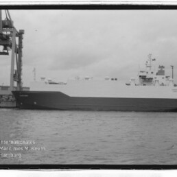 Großes weißes Ro‑Ro‑Frachtschiff am Kai unter bedecktem Himmel, links ein Hafenkran und unten links ein Museumswasserzeichen.