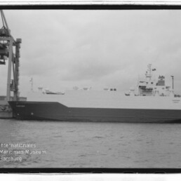 Großes Frachtschiff am Kai unter einem Hafenkran, schwarz-weiß Aufnahme mit Filmrand und Museumslogo unten links.