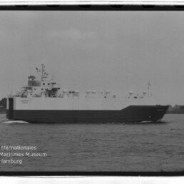 Schwarzweißfoto eines großen RoRo-Frachtschiffs auf ruhigem Wasser mit Ufer im Hintergrund und Museumslogo unten links.